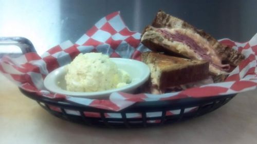 A basket with a grilled sandwich and a side of creamy potato salad, lined with red and white checkered paper.