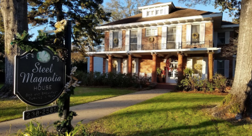 A charming brick house with a porch, decorated for the holidays, and a sign reading "Steel Magnolia House."