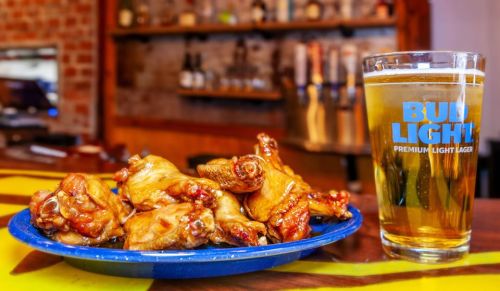 A plate of glazed chicken wings next to a glass of Bud Light beer on a colorful bar countertop.
