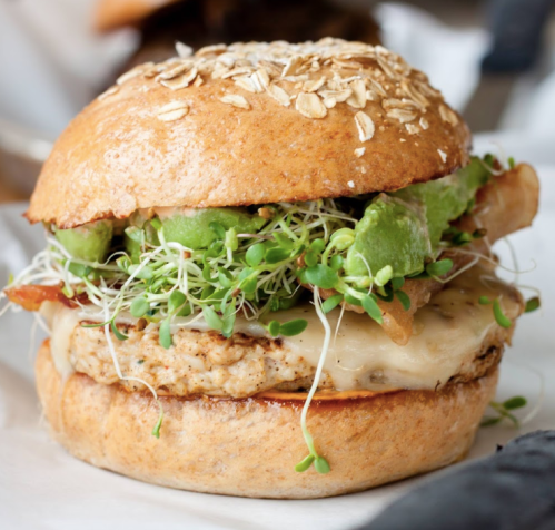 A hearty sandwich with a whole grain bun, filled with sprouts, avocado, cheese, and a chicken patty.