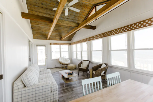 Bright sunroom with wooden beams, large windows, and cozy seating, overlooking a scenic view.