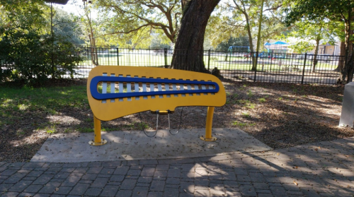 A large, yellow outdoor musical instrument resembling a xylophone, set in a park with trees and a playground in the background.
