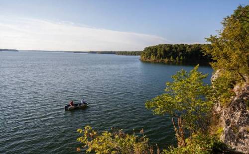 Two people in a small boat on a calm lake, surrounded by trees and a clear blue sky.