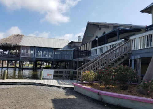 A waterfront building with a thatched roof, wooden stairs, and a garden near the water's edge on a sunny day.