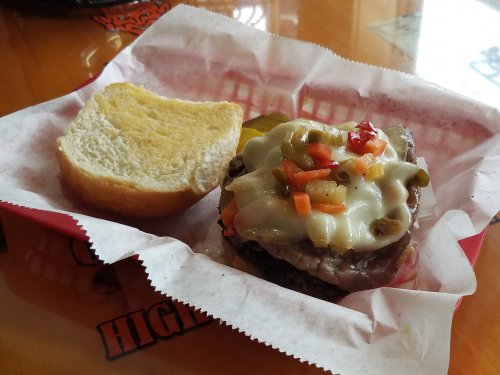 A sandwich with a beef patty topped with melted cheese, pickles, and diced vegetables, served with a slice of bread.
