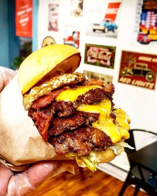 A towering burger stacked with multiple beef patties, cheese, bacon, and lettuce, held in a hand with a retro backdrop.