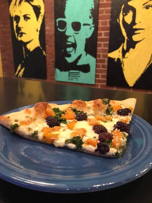 A slice of pizza topped with kale, blackberries, and cheese on a blue plate, with colorful portraits in the background.