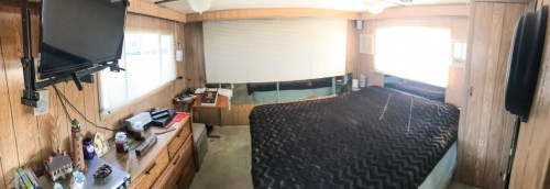 A cozy bedroom with wooden paneling, a large bed covered in a dark blanket, a TV, and a dresser with various items.