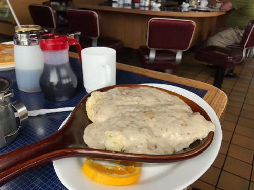 A plate of biscuits smothered in creamy sausage gravy, served with an orange slice, in a diner setting.