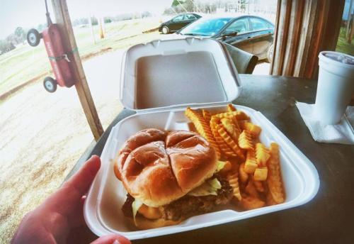 A hand holds a takeout container with a burger and crinkle-cut fries, with a car and grassy field in the background.