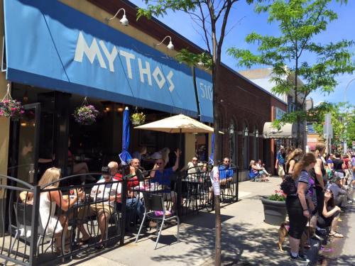 Outdoor dining scene at Mythos restaurant, with patrons enjoying meals on a sunny day, surrounded by greenery.
