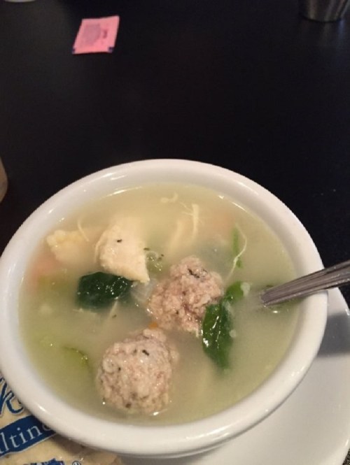 A bowl of soup with meatballs, vegetables, and greens in a clear broth, served with a spoon.
