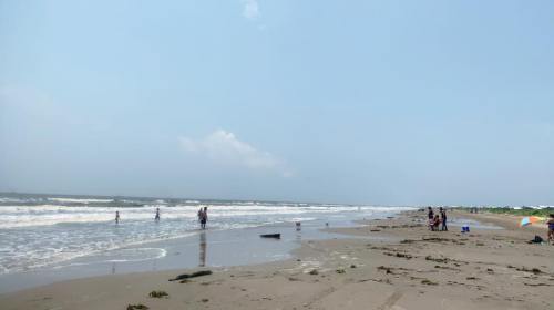 A sunny beach scene with people walking along the shore, waves crashing, and colorful umbrellas scattered on the sand.