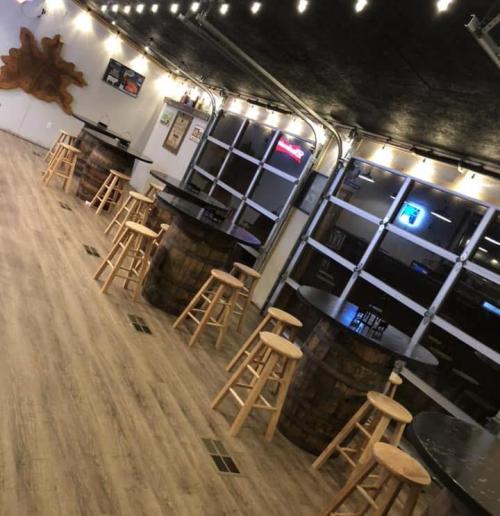 Interior of a rustic bar with wooden tables, stools, and string lights, featuring a garage door and wooden decor.