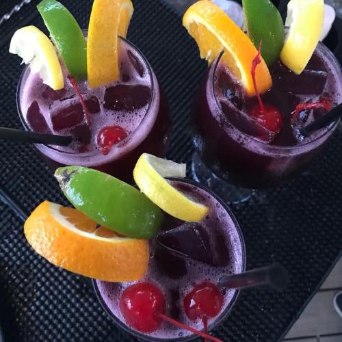 Three colorful cocktails with cherries, citrus slices, and ice, served in glasses on a textured surface.