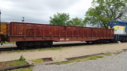 A red train flatcar parked on a gravel track, with a blue locomotive in the background and trees nearby.
