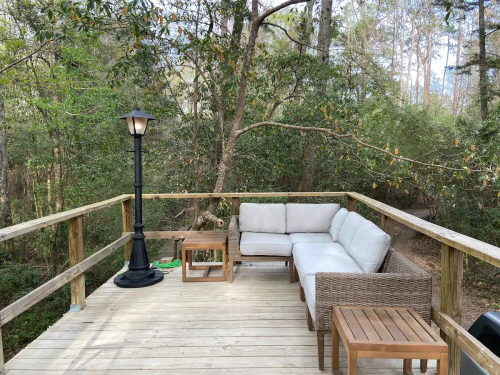 A cozy outdoor deck with a sectional sofa, wooden tables, and a lamp, surrounded by trees and greenery.