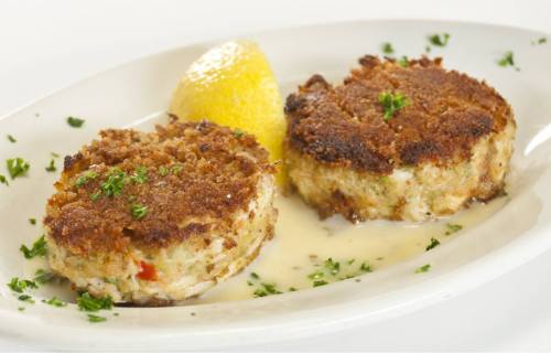 Two golden-brown crab cakes on a plate, garnished with parsley and served with a lemon wedge.