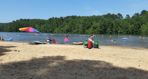 A sandy beach by a lake with people swimming, lounging under umbrellas, and enjoying a sunny day.