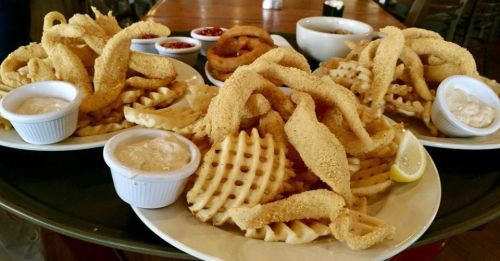 Two plates of golden fried calamari served with waffle fries, dipping sauces, and lemon wedges on a wooden table.