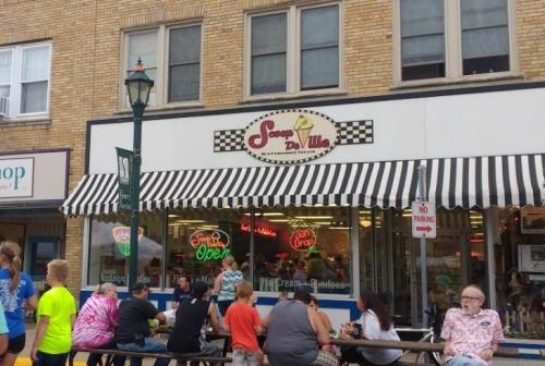 A busy ice cream shop with a striped awning, people sitting outside, and a bright "Open" sign in the window.