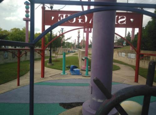 Colorful playground equipment with slides and pathways, surrounded by green grass and trees under a cloudy sky.