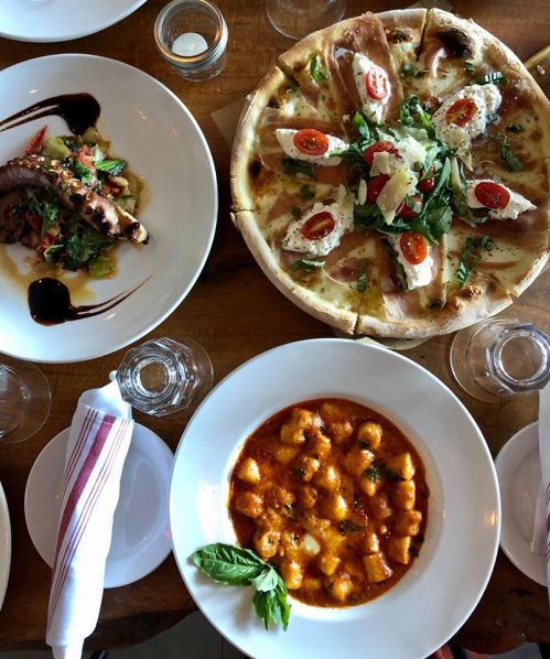 A table spread featuring a pizza with toppings, a plate of gnocchi in sauce, and a dish with grilled octopus.