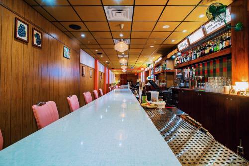 A retro diner interior featuring a long, light blue counter, pink chairs, and a well-stocked bar in warm wooden decor.