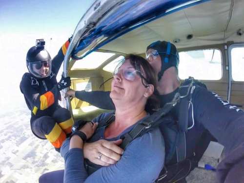 A woman prepares to skydive, seated in a plane with an instructor beside her and another skydiver at the open door.