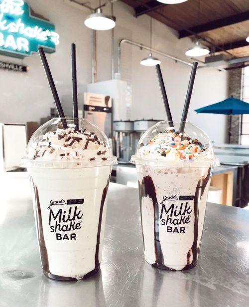Two milkshake cups topped with whipped cream, chocolate drizzle, and sprinkles, sitting on a counter in a bright café.