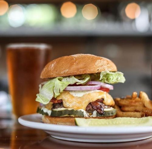A juicy burger with lettuce, tomato, onion, cheese, and pickles, served with fries and a glass of beer in the background.