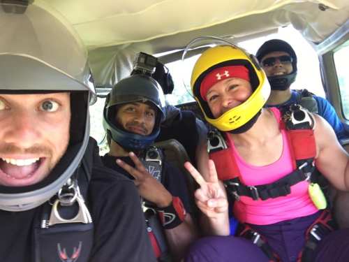 A group of four people in skydiving gear, smiling and posing for a selfie inside a plane.