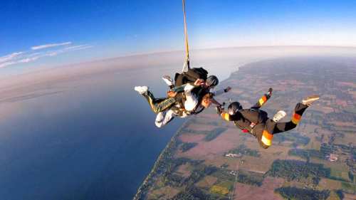 Three skydivers in freefall above a scenic landscape with water and fields below, under a clear blue sky.
