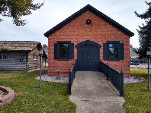 A brick building with black trim and a ramp, surrounded by grass and a log cabin in the background.