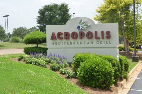 Sign for Acropolis Mediterranean Grill surrounded by green shrubs and flowers on a sunny day.