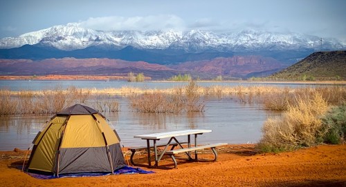 A tent by a lake with snowy mountains in the background and a picnic table nearby, surrounded by dry vegetation.