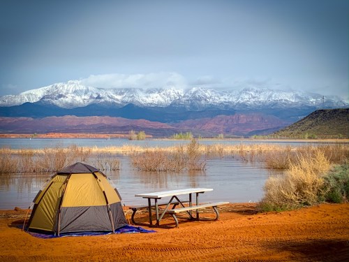 A tent and picnic table by a lake, with snow-capped mountains in the background and a clear sky.