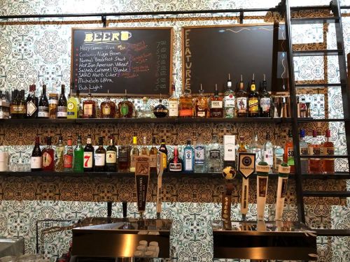 A bar shelf displaying various liquor bottles and taps, with a chalkboard menu listing beer options and features.