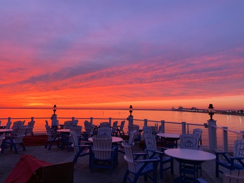A vibrant sunset over calm waters, with white chairs and tables lining a wooden deck.
