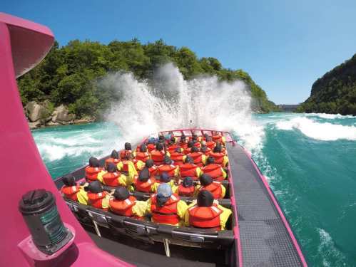 A group of people in bright life jackets on a boat, splashing through turquoise waters with waves crashing around them.