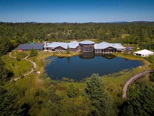 Aerial view of a modern building by a serene pond, surrounded by lush greenery and mountains in the background.