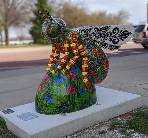 Colorful bee sculpture with intricate patterns, perched on a vibrant flower-covered mound in an outdoor setting.
