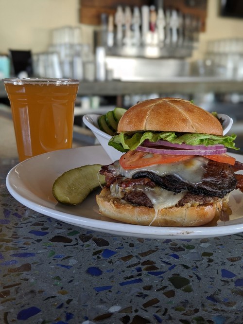 A juicy burger with lettuce, tomato, and cheese, served with a pickle and a glass of beer on a table.