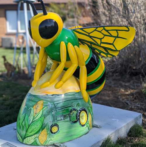 A colorful bee sculpture with yellow and green stripes, perched on a corn-themed base featuring a tractor design.