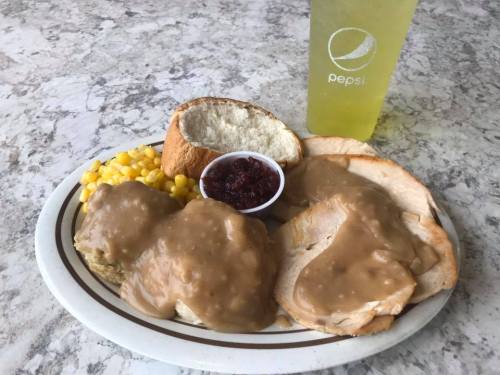 A plate with turkey and gravy, corn, a roll, and a small cup of cranberry sauce, alongside a Pepsi drink.