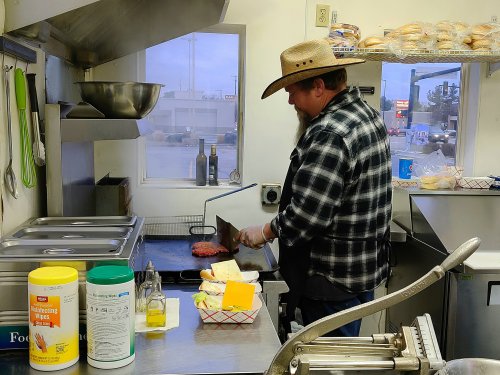 A man in a cowboy hat cooks on a grill in a diner kitchen, surrounded by condiments and food prep items.