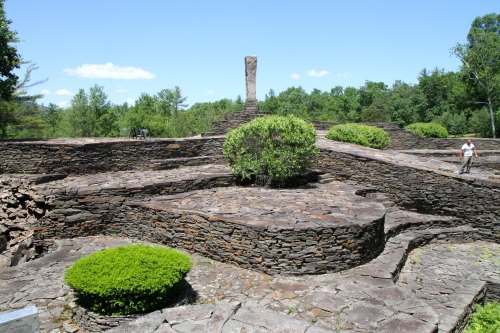 A stone structure with winding paths and greenery, set in a wooded area under a clear blue sky.