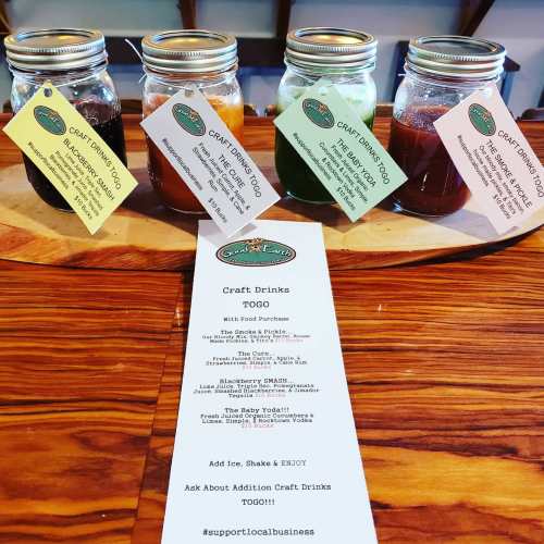 Four jars of craft drinks on a wooden platter, with a menu listing drink options and a note to support local business.