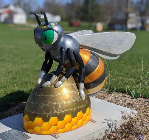 A colorful bee sculpture with large eyes and metallic wings perched on a honeycomb structure in a grassy area.