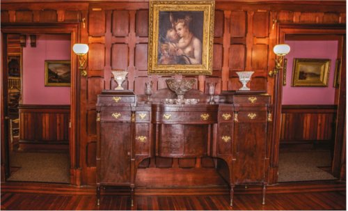 A wooden sideboard with decorative drawers and a painting above, set against a paneled wall in a warmly lit room.
